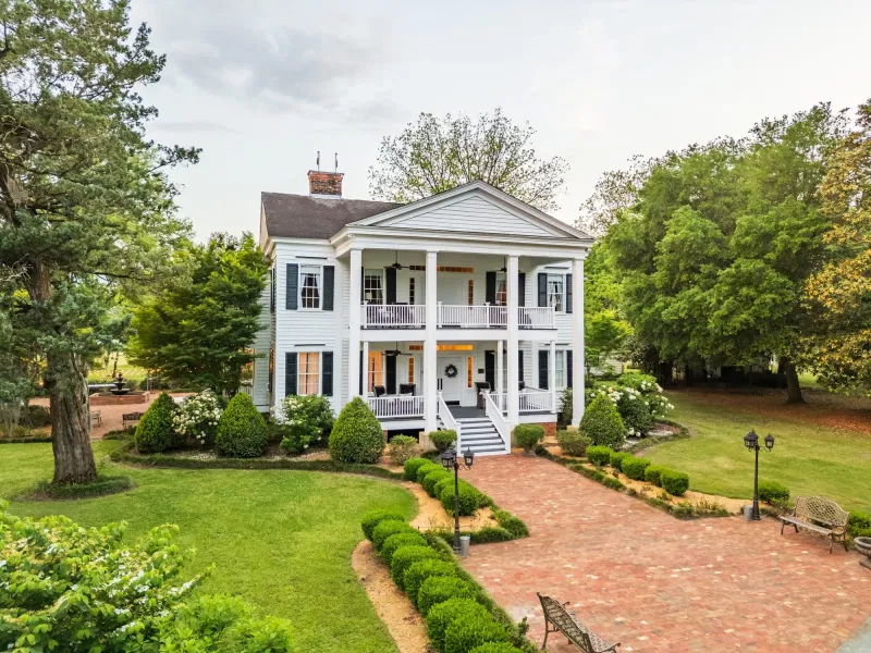 White Southern home with tall columns, a brick drive, and mature trees.